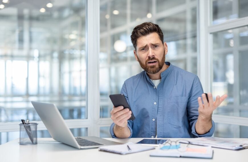 Upset and disappointed man with phone in hands. office worker at workplace looking anxiously at camera, working with laptop inside office at workplace